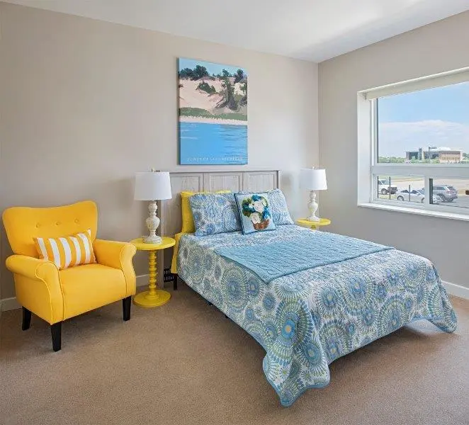 Memory care bed with blue covers, between a yellow chair and window, with a painting of a beach overhead